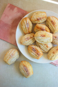 Many madeleines in a white plate, and two madeleines on the side. Pink napkin under the plate.