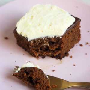 Portion of gingerbread cake with cream cheese frosting on a pink plate and golden fork with a piece of the cake.