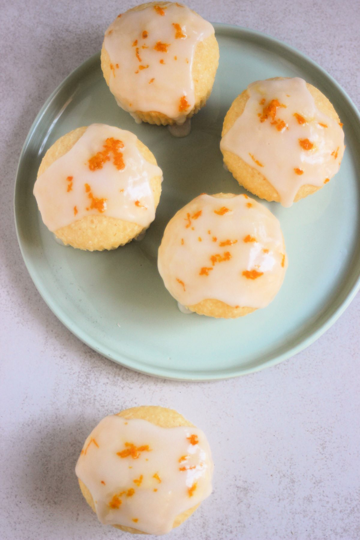 Orange coconut muffins with icing on an aqua-green plate and one muffins beside seen from above.