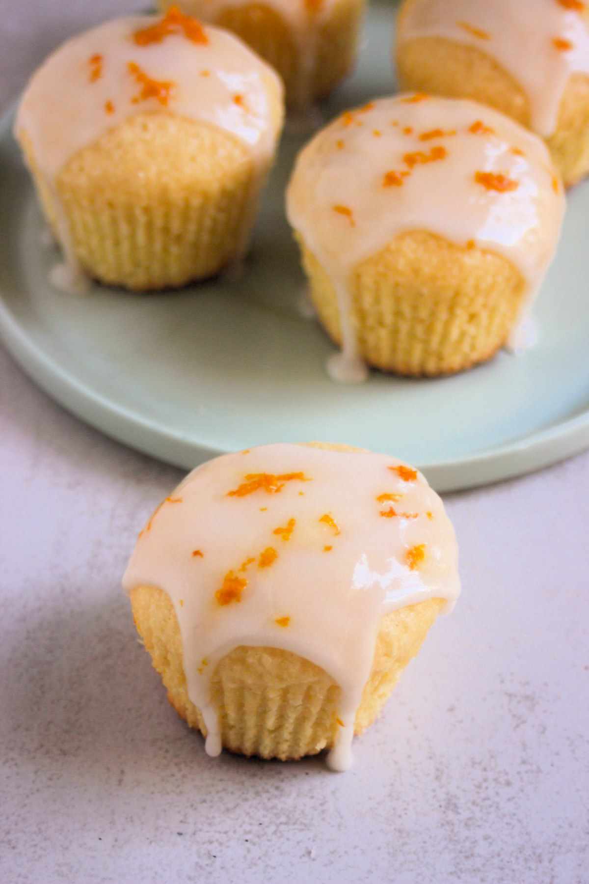 Orange coconut muffin with icing on a white surface. More muffin behind on an aqua-green plate.
