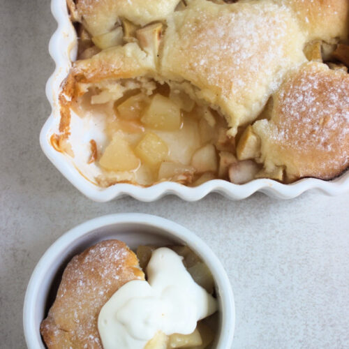 Rectangular white baking dish with pear cobbler without a portion and small bowl with pear cobbler with whipped cream seen from above.