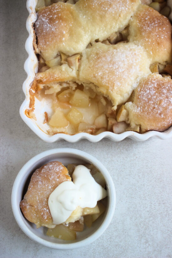 Rectangular white baking dish with pear cobbler without a portion and small bowl with pear cobbler with whipped cream seen from above.
