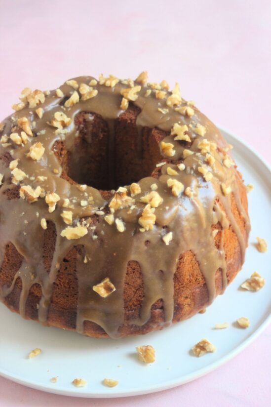Banana bundt cake with brown sugar icing and walnuts on a white plate. Pink surface.