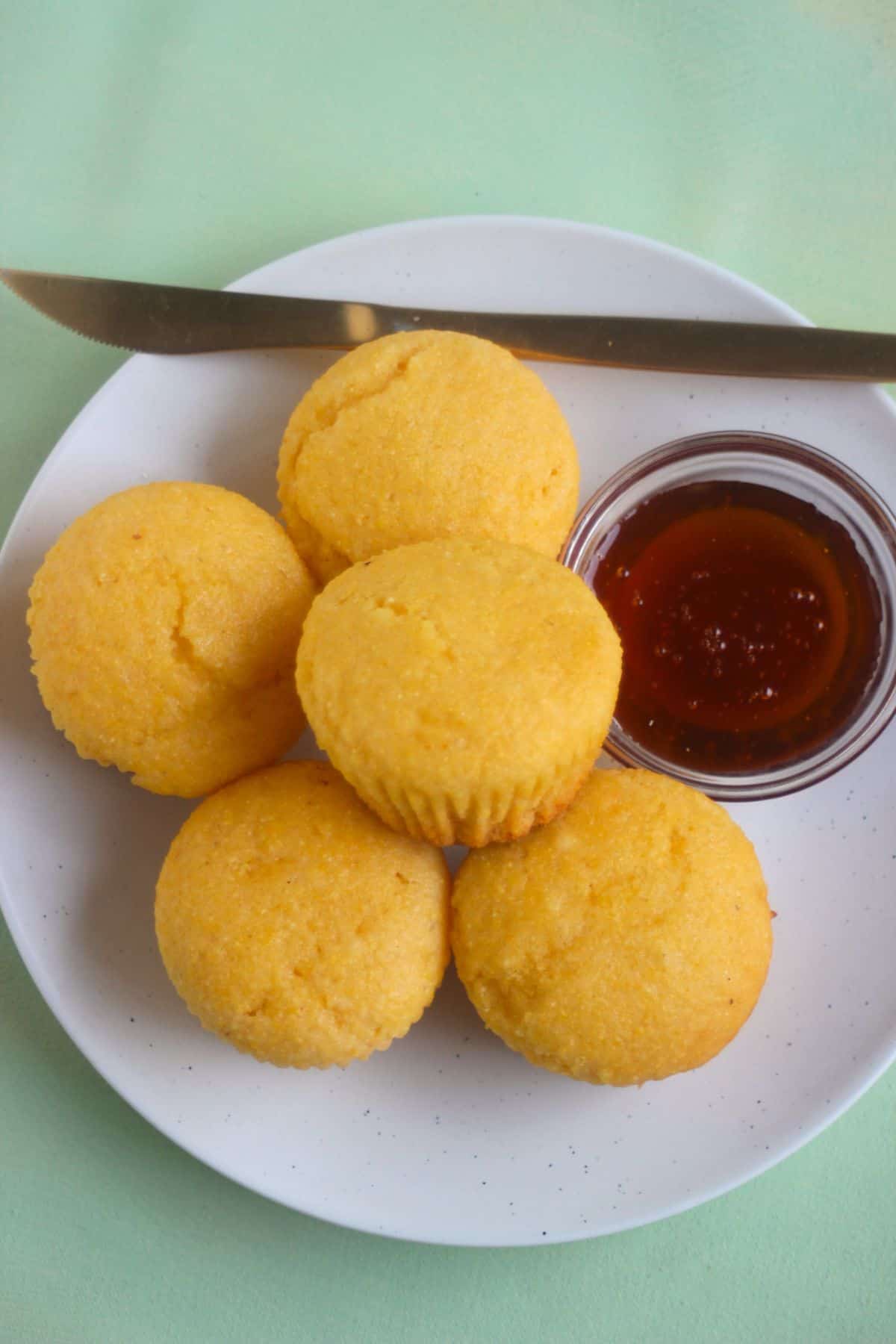 Cornbread muffins, a dip of honey, and a golden knife on a white plate seen from above.