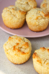 Two coconut muffins on a white surface. More muffins behind on a pink plate.