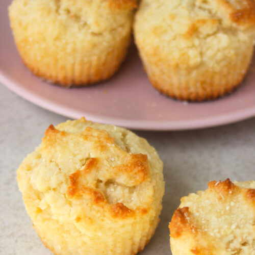 Two coconut muffins on a white surface. More muffins behind on a pink plate.