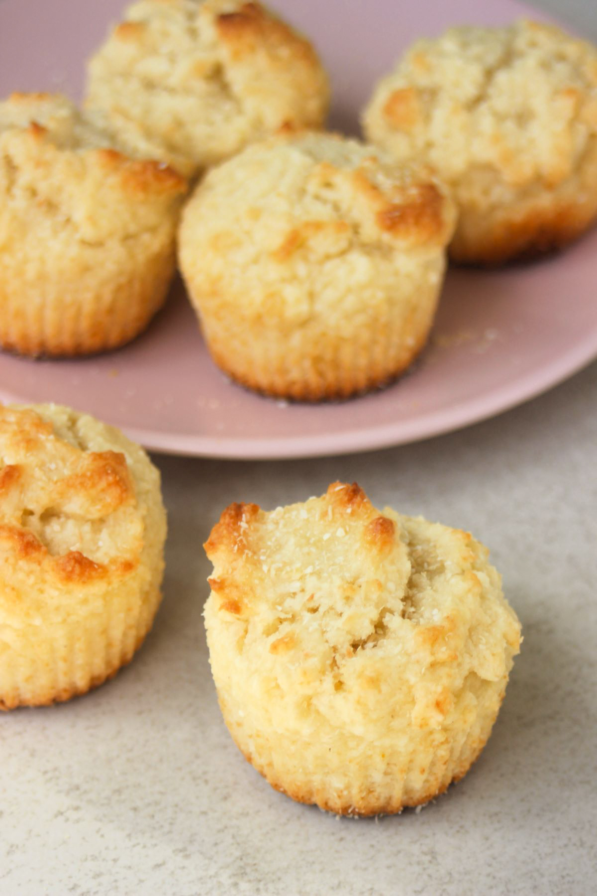 Two coconut muffins on a white surface. More muffins behind on a pink plate.