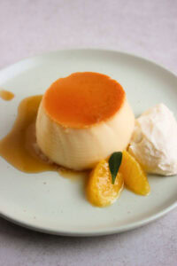 Orange flan with caramel, whipped cream, and fresh orange slices, on an aqua-green plate.