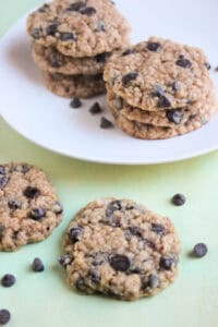 Oatmeal chocolate chip cookie on a light-green surface, chocolate chips scattered, and more cookies behind on a white plate.