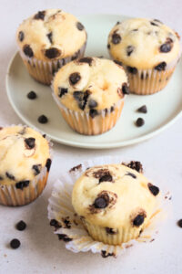 Chocolate chip muffin with the paper liner open on a white surface, and chocolate chips scattered beside. More muffins behind on an aqua-green plate.