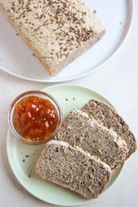 Small plate with three slices of buckwheat bread and a dip of jam seen from above. Beside a big white plate with a buckwheat bread loaf.