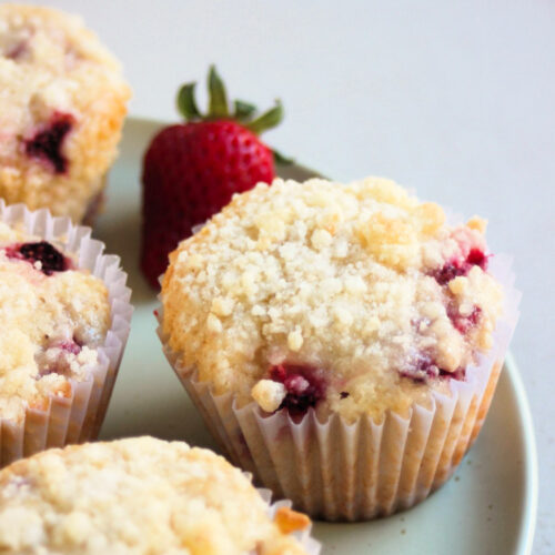 Strawberry streusel muffins with liners on an aqua green plate. A fresh strawberry.