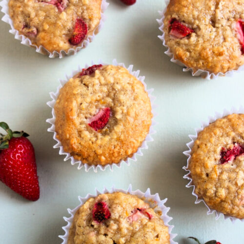 Strawberry oatmeal muffins with liners and fresh strawberries on a light blue surface seen from above.
