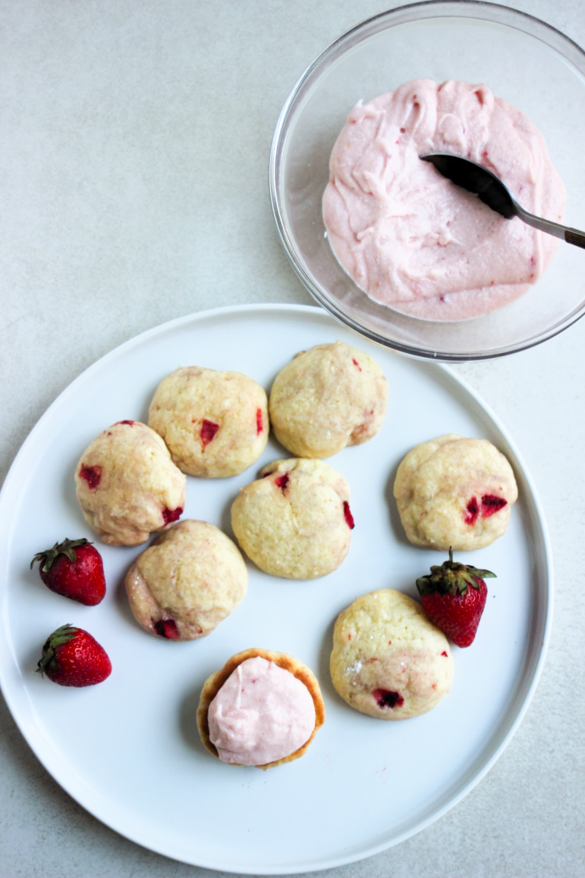 White plate with whoopie pie biscuits and fresh strawberries seen from above. One is topped with strawberry cream. Beside, a glass bowl with strawberry cream and a spoon.