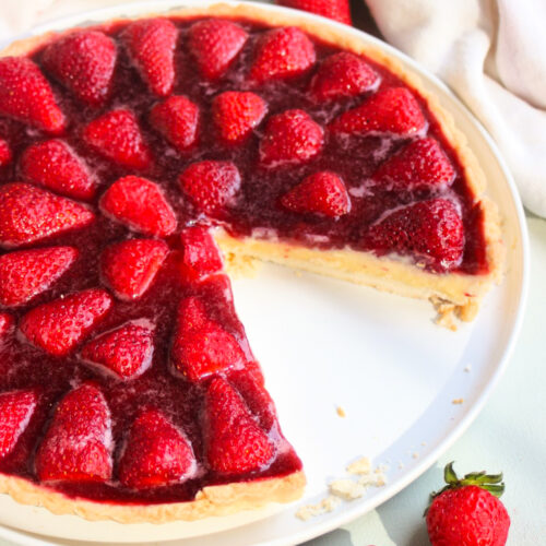 Partial strawberry tart on a white plate with a white kitchen towel and fresh strawberries. White surface.