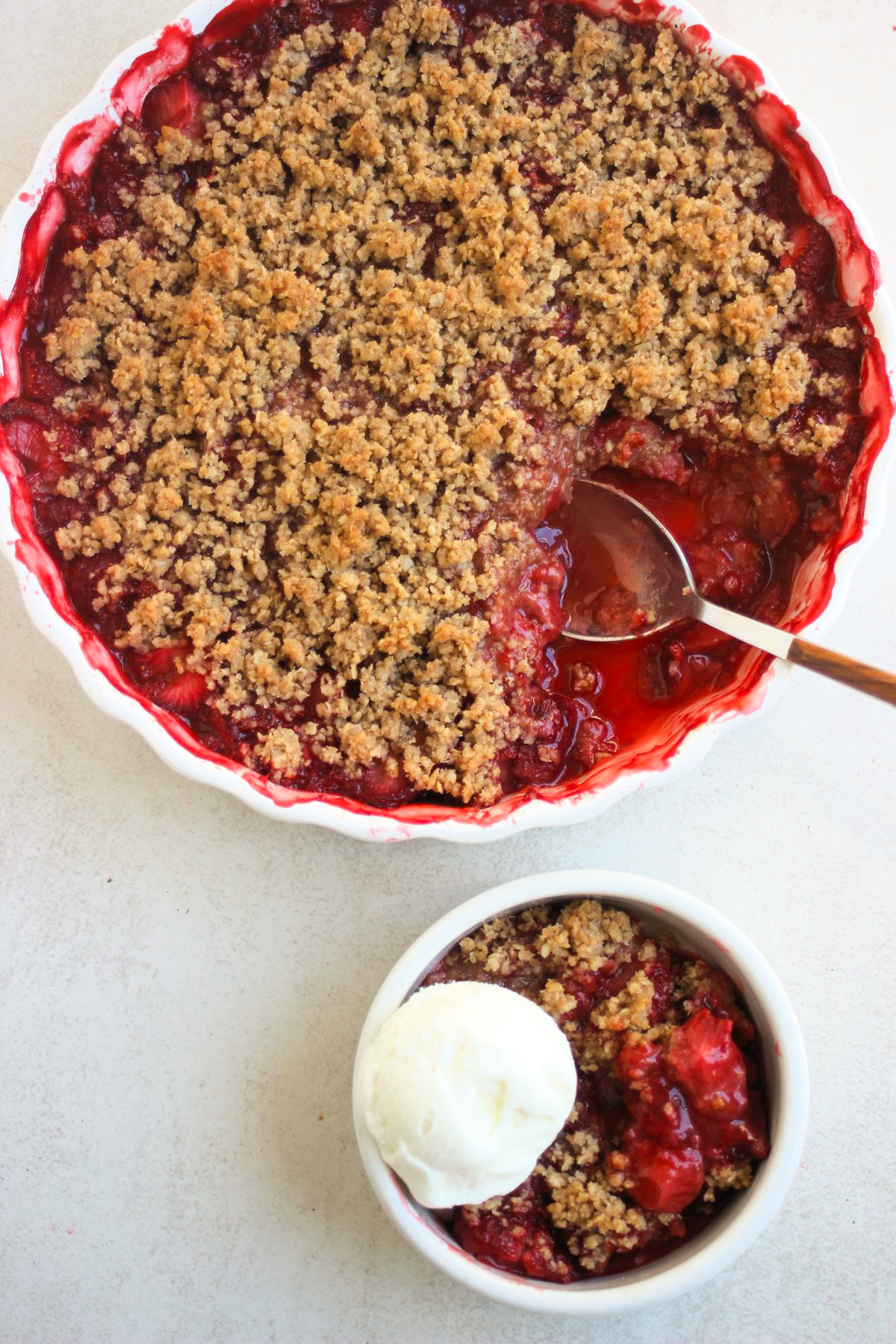 Top view of a white bowl with strawberry crisp and a scoop of ice cream and round baking dish with strawberry crisp and a spoon.