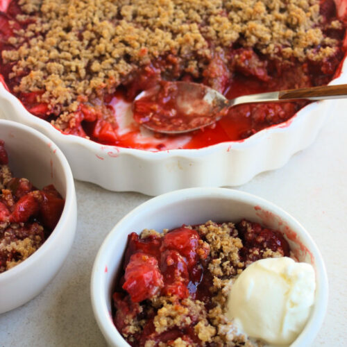White bowl with strawberry crisp and a scoop of ice cream. Behind, a round baking dish with strawberry crisp.