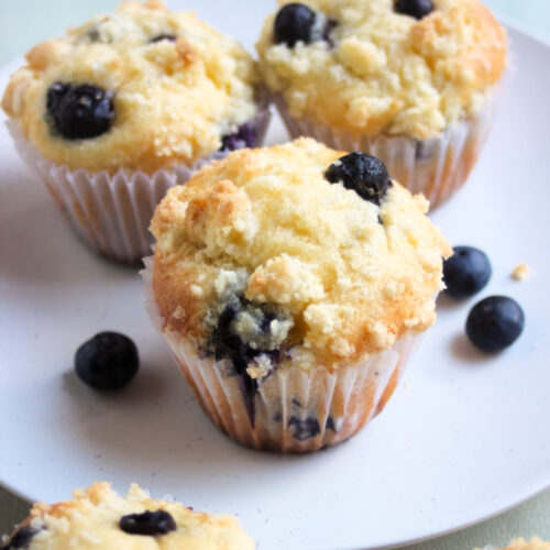 Blueberry streusel muffins in liners on a white plate. Fresh blueberries on the sides.