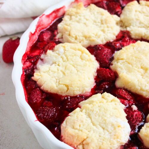 Partial view of strawberry cobbler on a round baking dish. White kitchen towel behind.