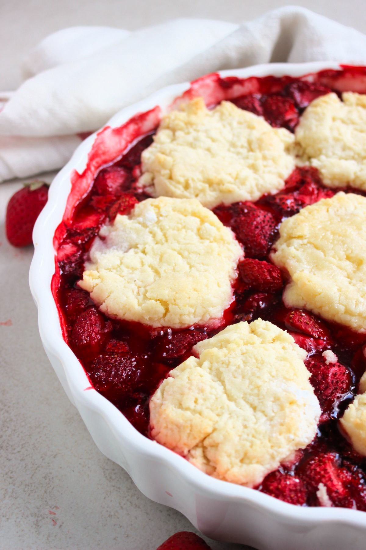 Partial view of strawberry cobbler on a round baking dish. White kitchen towel behind.