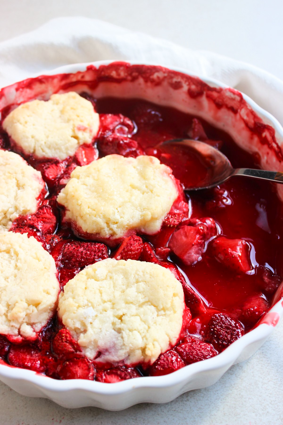 Partial view of strawberry cobbler, without some portions, and a spoon on a round baking dish. White kitchen towel behind.