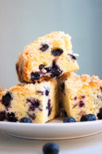 Squares of blueberry coffee cake on a white plate. One is above the others. Grey background.