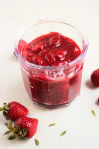 Jar with strawberry sauce on a white surface. Fresh strawberries and tiny leaves on the side.
