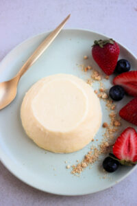 Top view of lemon panna cotta on an aqua green plate. A golden spoon, crumbs, fresh strawberries, and blueberries beside.