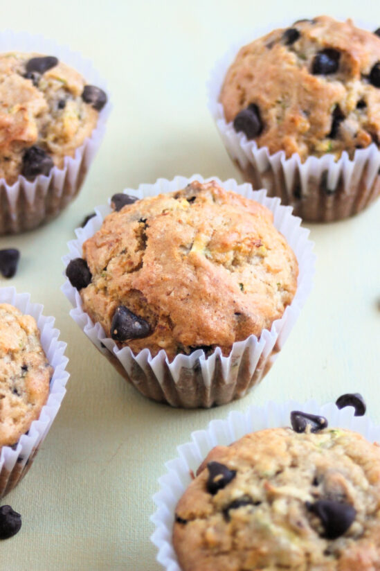 Zucchini chocolate chip muffins with liners on a light cream color surface.