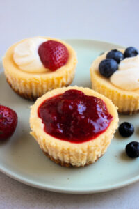 Mini cheesecakes on an aqua-green plate with different toppings: strawberry sauce, cream and strawberries, and cream and strawberry.