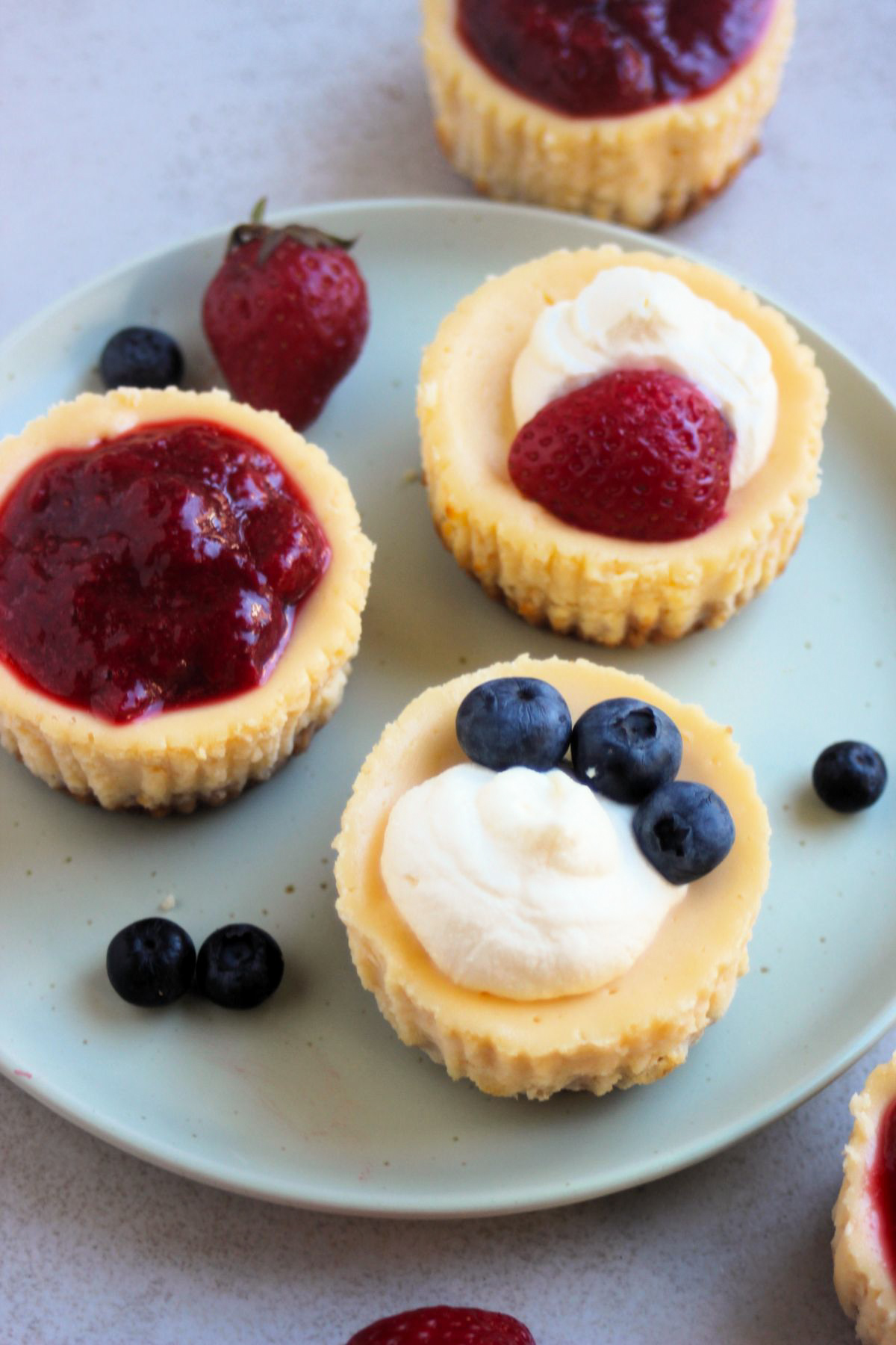 Mini cheesecakes on an aqua-green plate with different toppings: strawberry sauce, cream and strawberries, and cream and strawberry.
