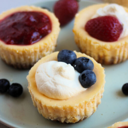 Mini cheesecakes on an aqua-green plate with different toppings: strawberry sauce, cream and strawberries, and cream and strawberry.