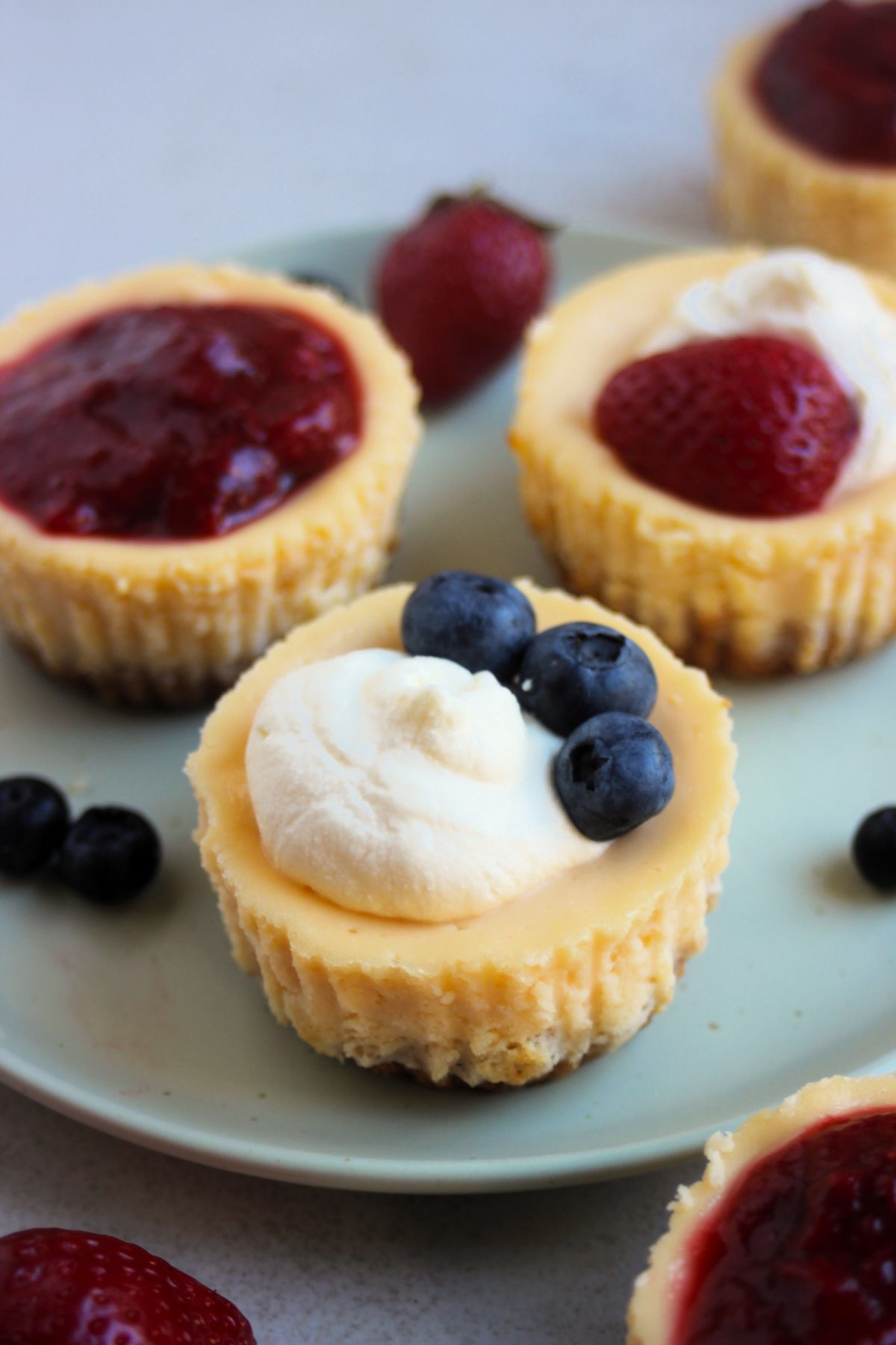 Mini cheesecakes on an aqua-green plate with different toppings: strawberry sauce, cream and strawberries, and cream and strawberry.