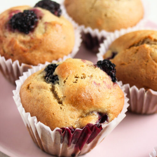 Four blackberry muffins with liners on a pink plate.
