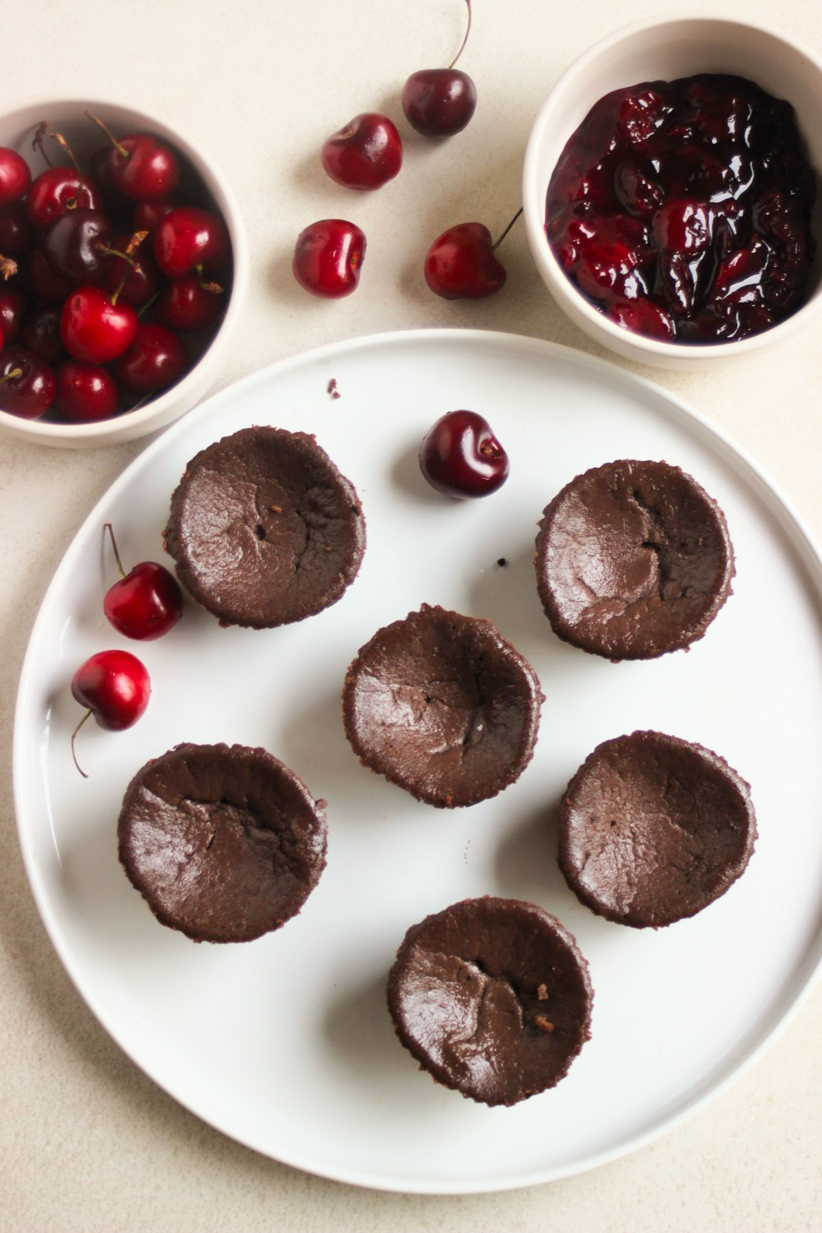 Top view of mini black forest cheesecakes without topping on a white plate. Two bowls, one with cherry pie filling, and the other with fresh cherries.