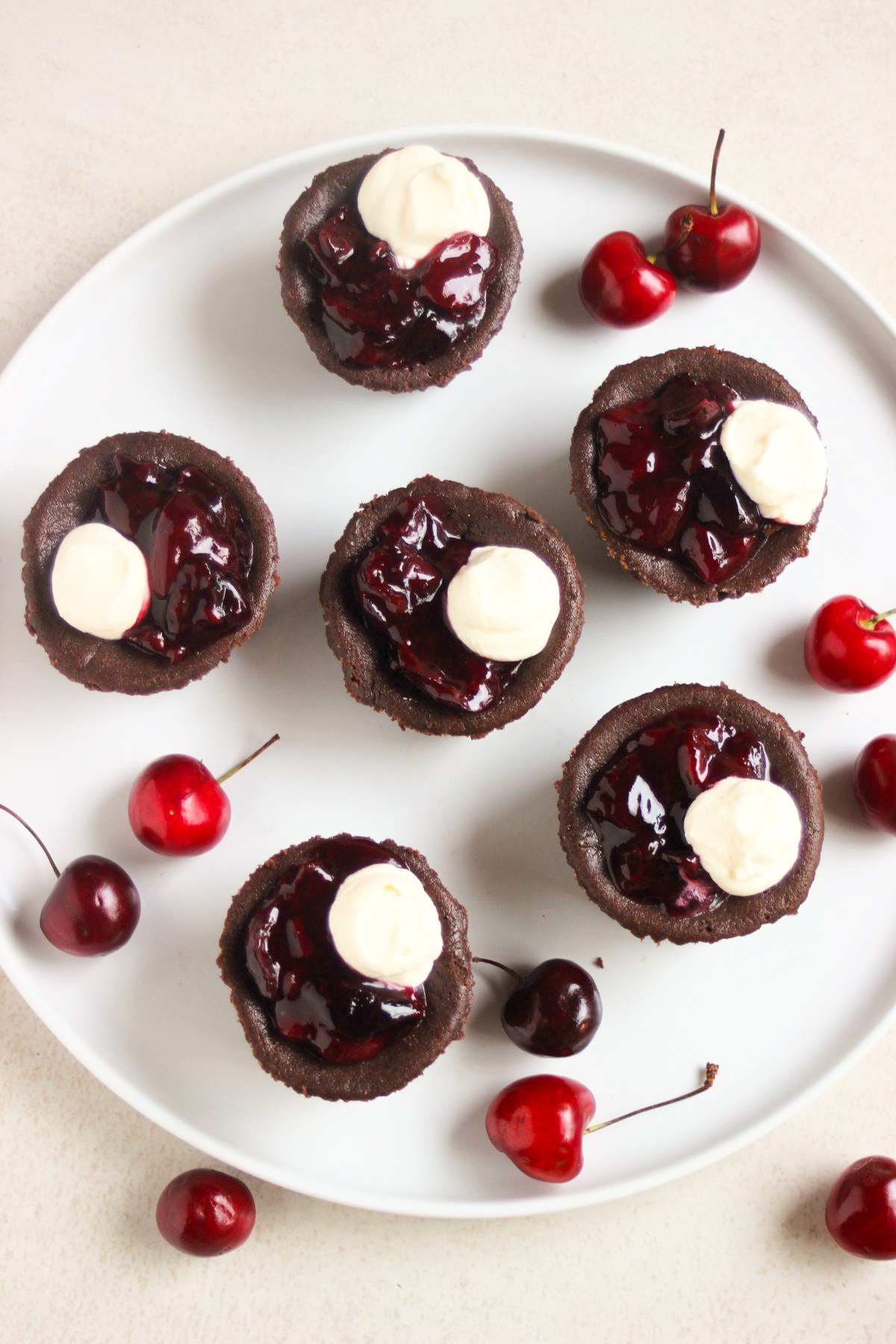 Top view of mini black forest cheesecakes with cherry topping and whipped cream on a white plate. Fresh cherries are scattered around.