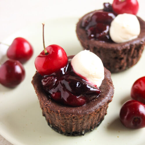 Two mini black forest cheesecakes with cherry topping, whipped cream, and fresh cherries on an aqua-green plate.