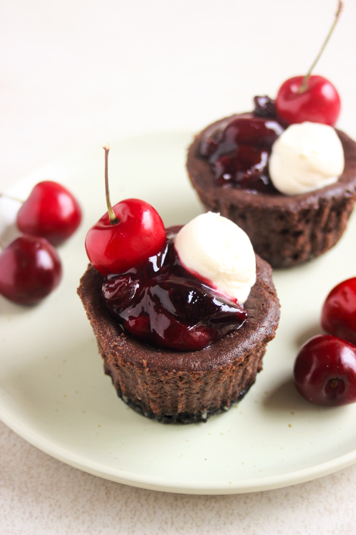 Two mini black forest cheesecakes with cherry topping, whipped cream, and fresh cherries on an aqua-green plate.