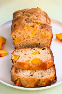 Peach loaf cake and slices of it, on a white plate. Fresh peach slices on the sides.