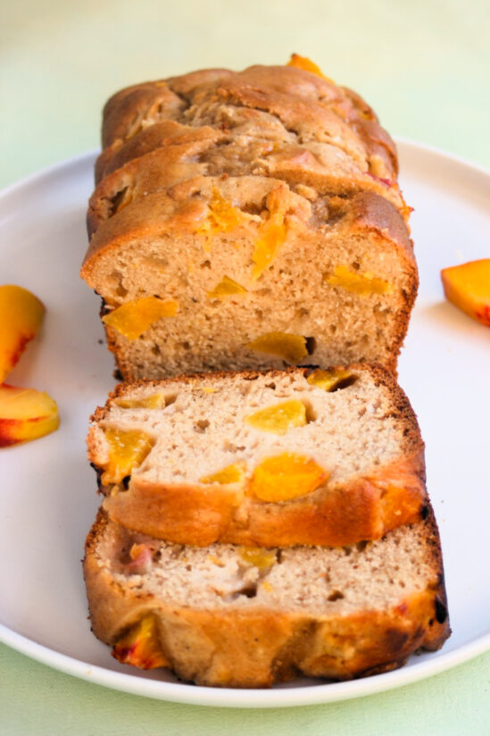 Peach loaf cake and slices of it, on a white plate. Fresh peach slices on the sides.