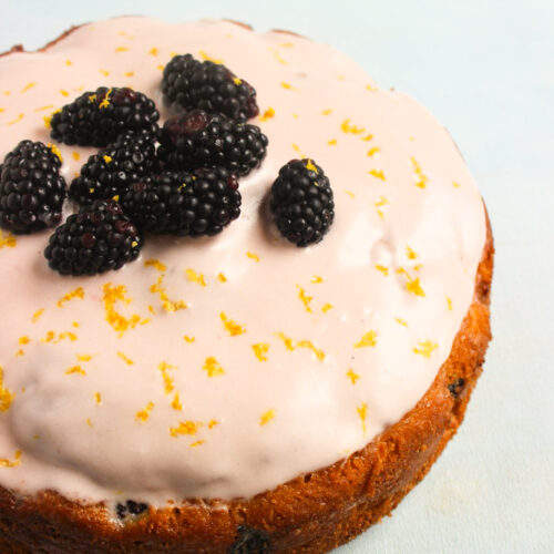 Blackberry cake with frosting and fresh blackberries on a light-blue surface.