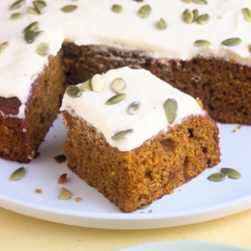 Portion of pumpkin cake with frosting and pumpkin seeds, and a pumpkin cake behind on a white plate.