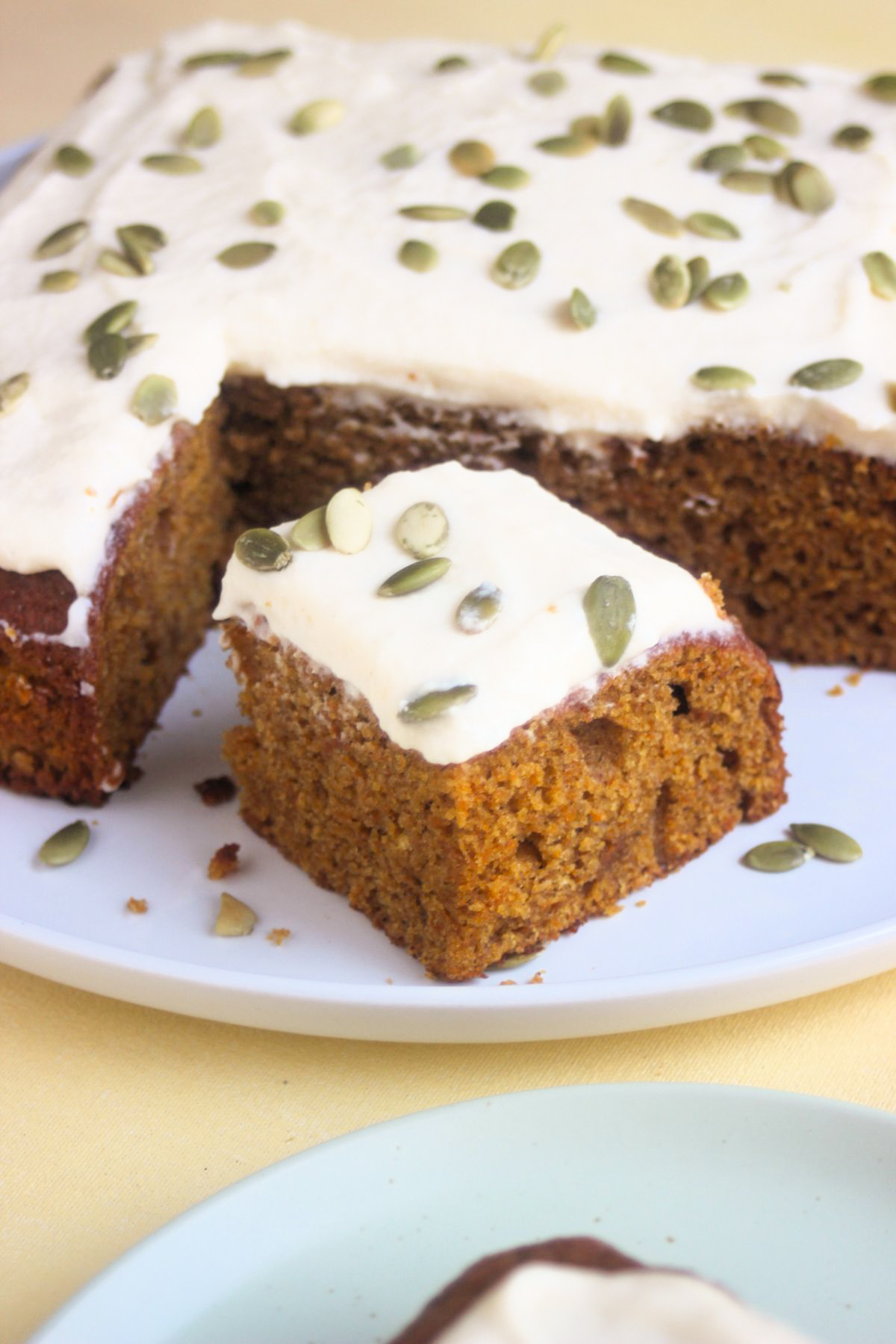 Portion of pumpkin cake with frosting and pumpkin seeds, and a pumpkin cake behind on a white plate.