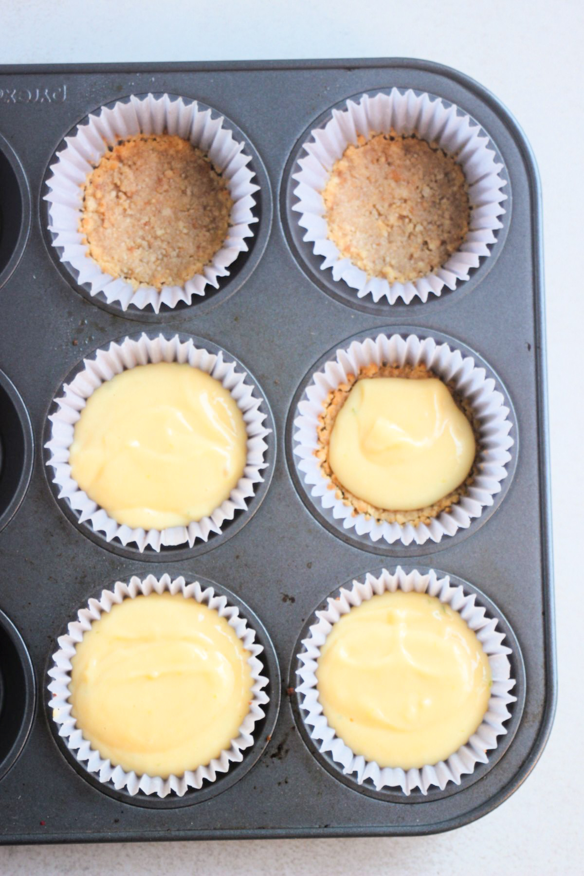 Top view of muffin pan with liners, four are filled with a light yellow mixture and two with cookie crust.