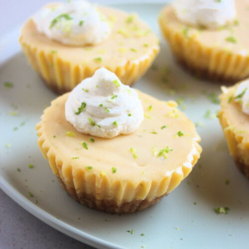 Mini key lime pie with whipped cream on an aqua-green plate. More mini cakes behind.