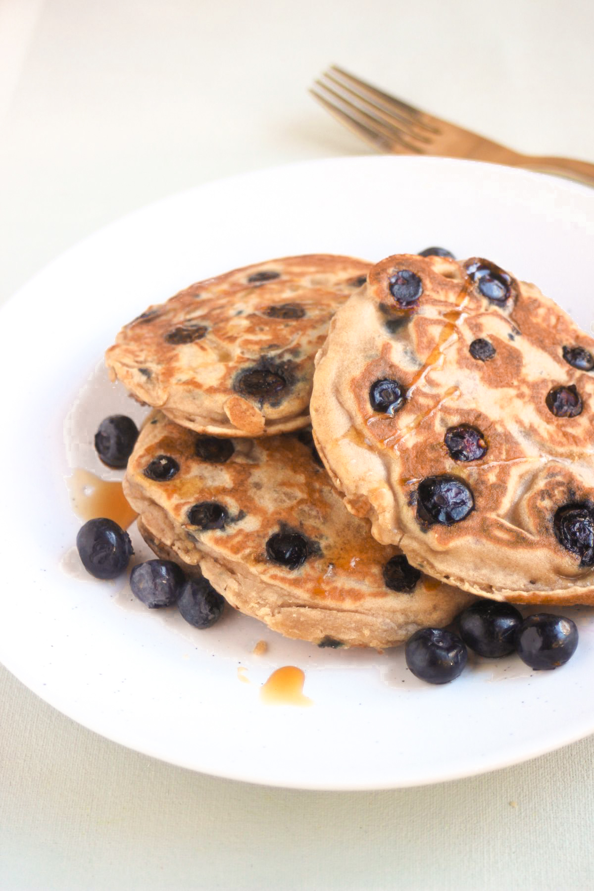 Three blueberry pancakes on a white plate and scattered blueberries. Golden fork behind the plate.