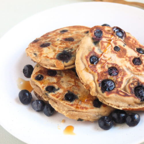 Three blueberry pancakes on a white plate and scattered blueberries. Golden fork behind the plate.