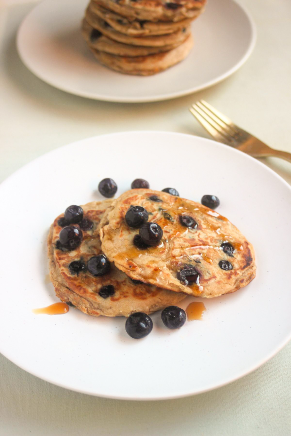 Two blueberry pancakes on a white plate with blueberries. Behind a golden fork and plate with stacked pancakes.
