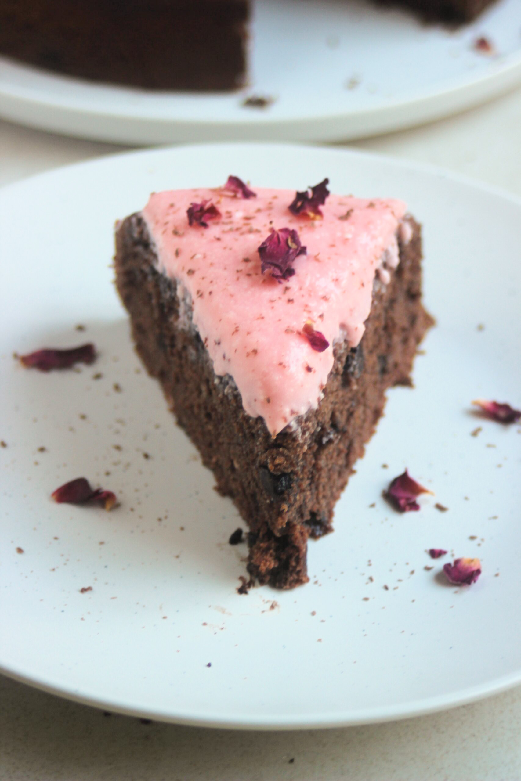 Portion of chocolate beet cake with pink frosting and rose leaves on a white plate.