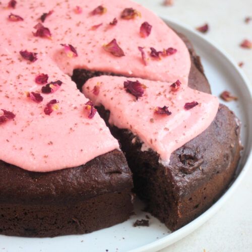 Chocolate beet cake with pink frosting and rose leaves on a white plate. A portion is cut off and sticks out of the cake.